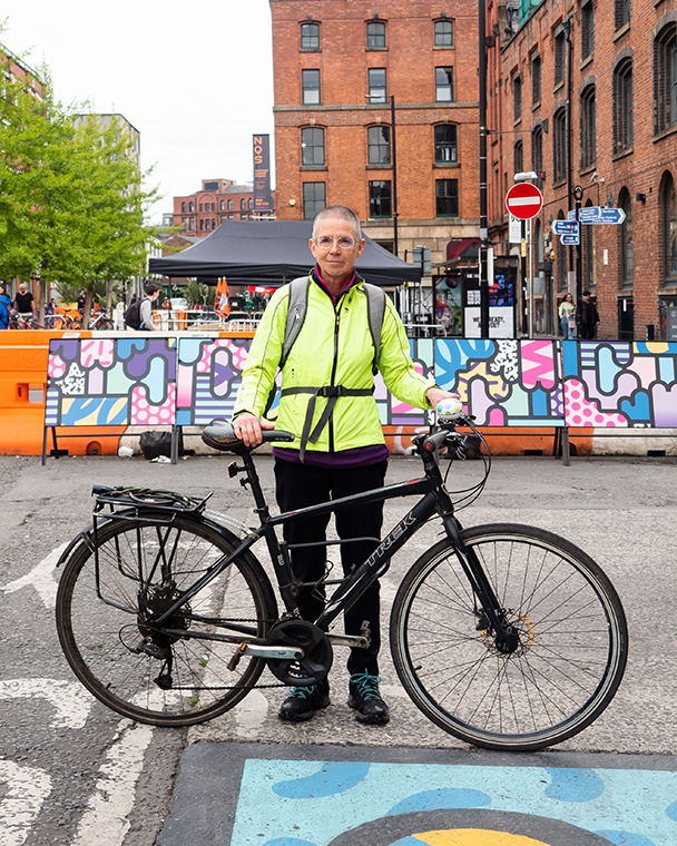 64-year-old Lizzie Gent from Manchester stands on a road in a florescent yellow jacket with her bike. Lizzie says: “It is important to acknowledge that older people have lived fascinating and interesting lives and have individual stories to tell.”