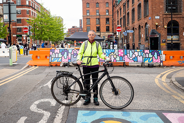 64-year-old Lizzie Gent from Manchester stands on a road in a florescent yellow jacket with her bike. Lizzie says: “It is important to acknowledge that older people have lived fascinating and interesting lives and have individual stories to tell.”